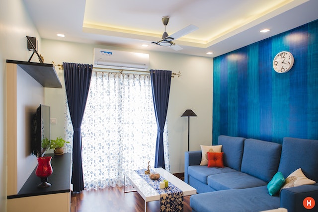 a living room with a blue accent wall in a rental property there is a blue sectional and a square white coffee table facing a television natural light streams in through the white curtains and lights up the space
